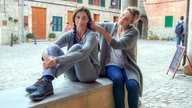 Filmszene aus dem Film "Insel der Zitronenblüten": Marina (gespielt von Elia Galera) und Anna (gespielt von Eva Martin) sitzen auf einem Mauervorsprung. Anna flechtet Marinas Haare. 