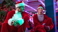 Eine Szene des Films "Jingle Bell Heist": Nick, gespielt von Connor Swindells, und Sophia, gespielt von Olivia Holt, sitzen in Weihnachtsmannkostümen auf dem Rand einer Tribüne. Die Kostümbärte haben sie heruntergezogen, er schaut sie interessiert an.