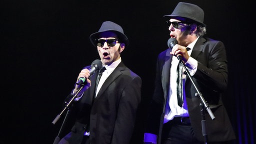 Florian Bender und Gregor Eckert als Elwood und Jake Blues auf der Bühne. Beide tragen schwarze Anzüge mit schwarzer Krawatte, Sonnenbrille und Hut. Beide stehen an Mikrofonen und singen. 