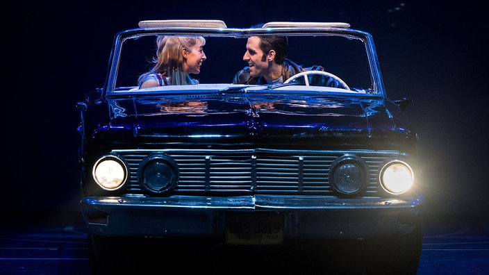 Darsteller Antonia Kalinowski und Philipp Büttner als Sandy und Danny auf der Bühne. Sie sitzen in einem Auto, er am Steuer, sie auf dem Beifahrersitz, und schauen einander lächelnd an.