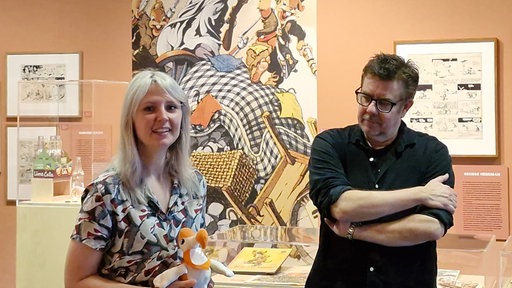 Eine Frau hält ein Ausstellungsstück in den Händen und lächelt in die Kamera. Neben ihr steht ein Mann und betrachtet das Ausstellungsstück. Im Hintergrund ist ein Gemälde und ein Glaskasten mit einem weiteren Ausstellungsstück.