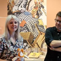 Eine Frau hält ein Ausstellungsstück in den Händen und lächelt in die Kamera. Neben ihr steht ein Mann und betrachtet das Ausstellungsstück. Im Hintergrund ist ein Gemälde und ein Glaskasten mit einem weiteren Ausstellungsstück.