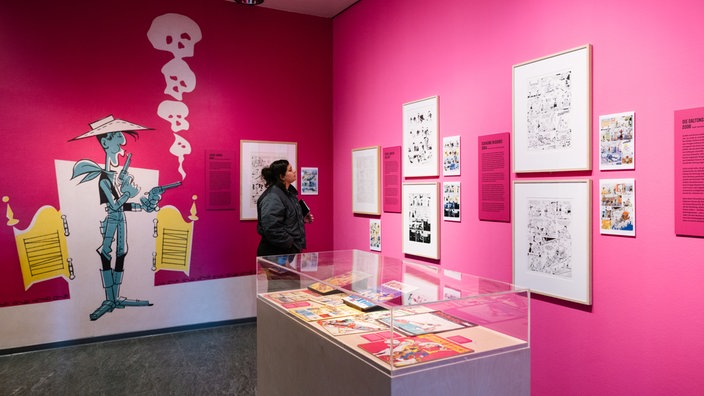 Ein Ausstellungsraum mit pinkfarbenen Wänden: Eine Besucherin schaut sich einen der Comics an, die in Rahmen an den Wänden hängen. Im Vordergrund ist ein Ausstellungskasten aus Glas mit "Lucky Luke"-Ausgaben zu sehen. 