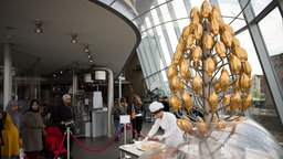 Der Schokobrunnen im Kölner Schokoladenmuseum: Die Elemente des Brunnens sind wie eine goldene Kakaobohne geformt. Neben dem Brunnen steht eine Mitarbeiterin, im Hintergrund sind Besucher zu sehen, die sich den Brunnen anschauen. 