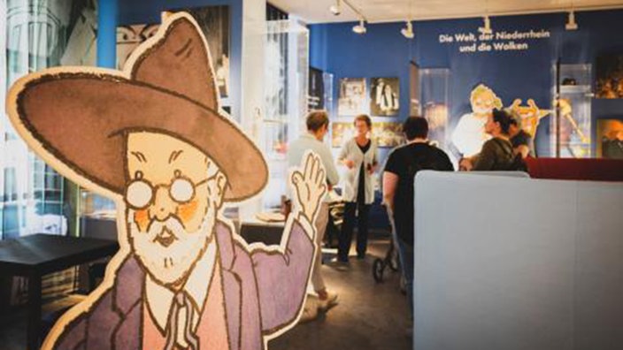 Dauerausstellung im Alten Landratsamt in Moers: Im Vordergrund des Bildes befindet sich ein illustrierter Aufsteller von Hanns-Dieter Hüsch, im Hintergrund sieht man die Ausstellung und einige Besucher. 