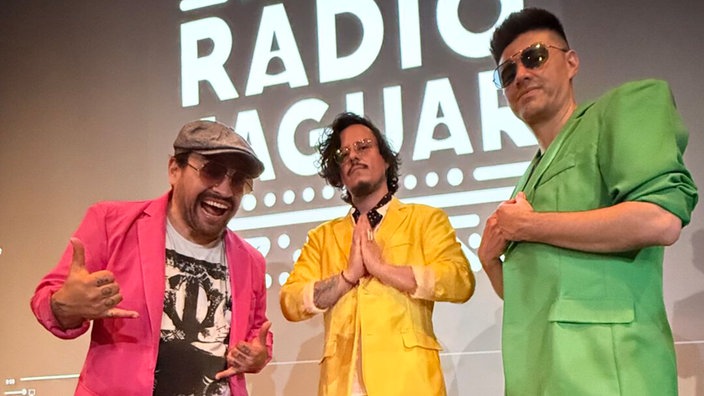 Die Mitglieder von "Radio Jaguar" in leuchtenden Sakkos in pink, gelb und grün vor einem schwarz-weißen Radio Jaguar Schriftzug. 