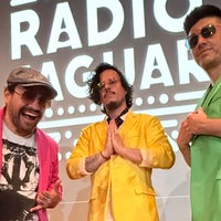 Die Mitglieder von "Radio Jaguar" in leuchtenden Sakkos in pink, gelb und grün vor einem schwarz-weißen Radio Jaguar Schriftzug. 