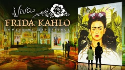 Das Bild zeigt eine farbenfrohe Ausstellung mit vielen Gemälden Frida Kahlos. Im Vordergrund rechts im Bild ist ein Selbstporträt von ihr zu sehen, davor stehen einige Besucher. Die Aufschrift "Viva Frida Kahlo, immersive Experience" ist außerdem zu sehen