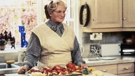 Eine Szene aus dem Film "Mrs. Doubtfire": Robin Williams steht als ältere Dame Mrs Doubtfire verkleidet in der Küche. 