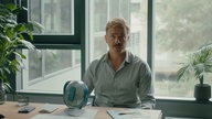 Szene aus dem Film " Pfau – Bin ich echt?": Matthias, gespielt von Albrecht Schuch sitzt an einem Schreibtisch. Hinter ihm befindet sich eine Fensterfront. 
