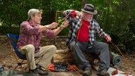 Szene des Films "Picknick mit Bären": Robert Redford als Bill Bryson und Nick Nolte als Stephen Katz sitzen auf Campingstühlen und stecken Zeltstangen zusammen.