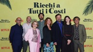 Ein Gruppenfoto des Casts des Films "Reich um jeden Preis" bei der Premiere auf dem roten Teppich. Die acht Schauspieler lächeln freundlich in die Kamera. Hinter ihnen an der gelben Wand steht der italienische Originaltitel "Ricchi a Tutti i costi".