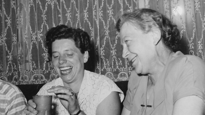 60er Jahre: Zwei Frauen sitzen lachend auf dem Sofa, eine von ihnen hält einen Würfelbecher in der Hand