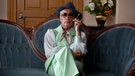 Eine Szene des Films "Swan Song": Udo Kier sitzt in extravaganter Kleidung und mit Zigarette in der Hand auf einem verschnörkelten Sofa