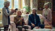 Eine Szene des Films "The Thursday Murder Club": Zwei ältere Herren und zwei ältere Damen in einem Wohnzimmer um ein Sofa versammelt