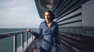 Eine Szene aus dem Film "The Woman in Cabin 10": Journalistin Laura "Lo" Blacklock, gespielt von Keira Knightley, steht an der Reling eines Kreuzfahrtschiffes. Sie trägt ein blaues Businesskleid und schaut ernst in die Ferne. 