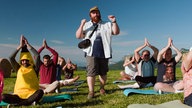 Szene aus dem Film "Was ist schon normal?": Personen beim Yoga auf einer grünen Wiese, in der Mitte steht ein Mann