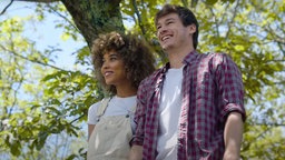 Szene aus dem Film " Zum Mars oder zu dir?": Daisy, gespielt von Alexandra Schipp, steht neben Alex, gespielt von Kyle Allen unter einem Baum. Durch die Baumkrone ist ein blauer Himmel zu sehen, beide scheinen auf etwas hinauszublicken und lächeln.