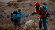 Eine Frau reicht einem Mann beim Wandern die Hände. Sie steht etwas höher auf einem Stein und hilft ihm nachzukommen.