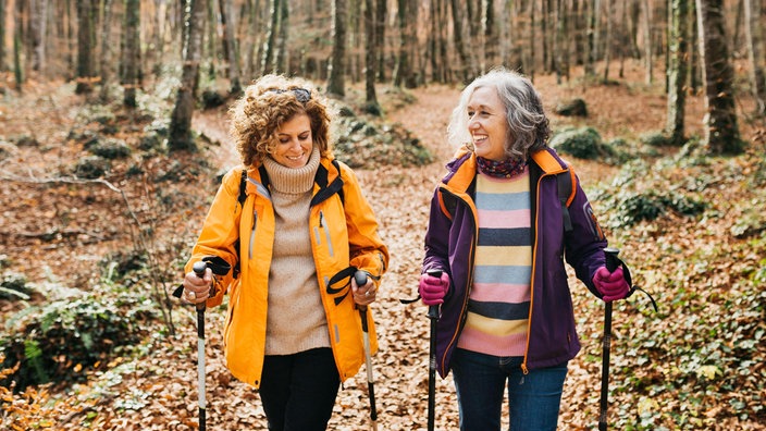 Zwei Freundinnen wandern im Wald. Sie sind einander zugewandt und lächeln. Beide tragen Wanderstöcke und praktische Kleidung.