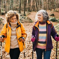 Zwei Freundinnen wandern im Wald. Sie sind einander zugewandt und lächeln. Beide tragen Wanderstöcke und praktische Kleidung.