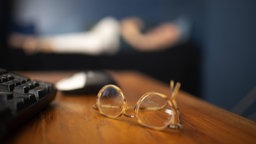 Im Vordergrund liegen auf einem Holztisch eine Brille, Maus und Tastatur, während man im Hintergrund eine schlafende Person erkennen kann