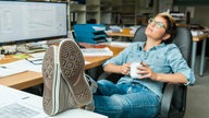 Eine Frau mit kurzen Haaren sitzt mit hochgelegten Beinen an einem Schreibtisch im Büro. Sie schaut entspannt in die Luft. In den Händen hält sie eine Kaffeetasse. 