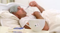 Ein Mann liegt im Bett mit einem Waschlappen auf der Stirn und einem Fieberthermometer im Mund. Im unteren Bildrand ist zu sehen, dass auf dem Nachttisch eine Packung Taschentücher liegt und eine Tasse Tee steht.