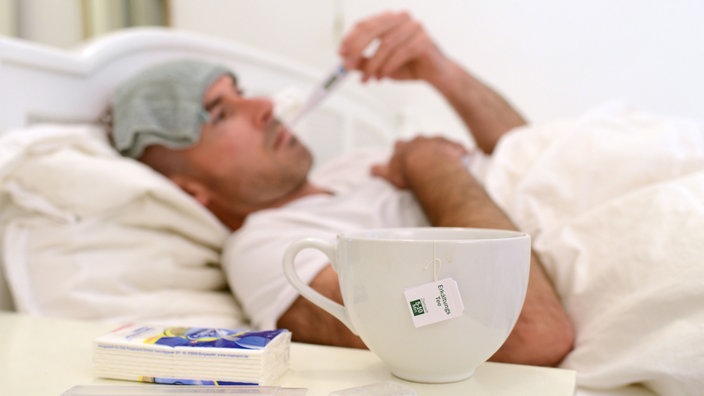 Ein Mann liegt im Bett mit einem Waschlappen auf der Stirn und einem Fieberthermometer im Mund. Im unteren Bildrand ist zu sehen, dass auf dem Nachttisch eine Packung Taschentücher liegt und eine Tasse Tee steht.