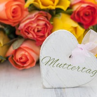 An einem Strauß mit roten, gelben und orangenen Rosen hängt ein Wweißes Herz, auf dem mit verschlungener Schrift "Muttertag" steht.