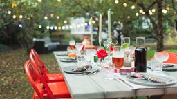Ein einladend gedeckter Tisch in einem Garten. Im Hintergrund hängen Lichterketten in den Bäumen.