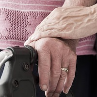 Nahaufnahme von Händen und dem Griff eines Rollators: Eine jüngere Frau unterstützt eine ältere Frau beim Gehen. 