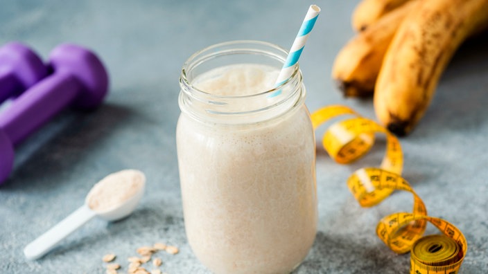 Ein Proteinshake in einem großen Glas mit einem blau-weißen Strohhalm. Daneben liegt ein Löffel mit Proteinpulver, zwei kleine, lila Hanteln, ein Bund Bananen und ein Maßband. 