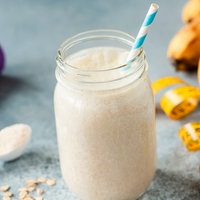 Ein Proteinshake in einem großen Glas mit einem blau-weißen Strohhalm. Daneben liegt ein Löffel mit Proteinpulver, zwei kleine, lila Hanteln, ein Bund Bananen und ein Maßband. 