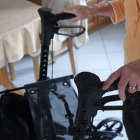 Eine ältere Frau läuft in ihrer Küche am Rollator. Ihre Hände an den Griffen sind im Fokus des Bildes, im Hintergrund sieht man ihren Küchentisch.