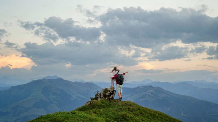 Ein Vater umarmt seine Tochter auf einer Bergspitze unter bewölktem Himmel und zeigt mit seiner Hand in die Ferne.