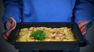 Eine schwarze Auflaufform wird mit Topflappen in die Kamera gehalten. Darin sind die gegarten Kartoffeln, verziert mit Kräutern.