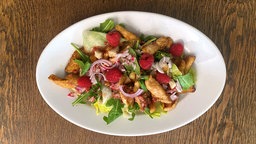 Blattsalat mit Hähnchenbruststreifen auf einem Teller, darauf ein paar Himbeeren