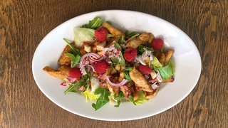 Blattsalat mit Hähnchenbruststreifen auf einem Teller, darauf ein paar Himbeeren