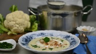 Angerichteter Suppenteller mit Blumenkohlsuppe, die mit Mandelblättern und Kräutern dekoriert ist, im Hintergrund ein Blumenkohl und Küchenutensilien