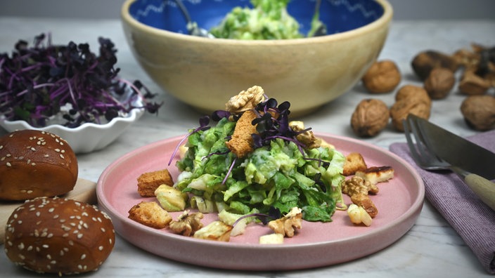 Endiviensalat mit Kartoffeldressing ist auf einem Teller angerichtet, daneben liegen Besteck, Laugenbrötchen und Walnüsse und im Hintergrund stehen zwei Schüsseln mit Salat