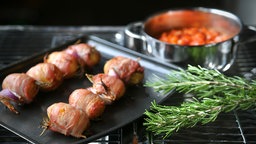 Kartoffelspieße mit Speck ummantelt auf einem schwarzen Blech. Danaben ein Rosmarinzweig. Im Hintergrund ein Topf gefüllt mit Paprika-Dip.