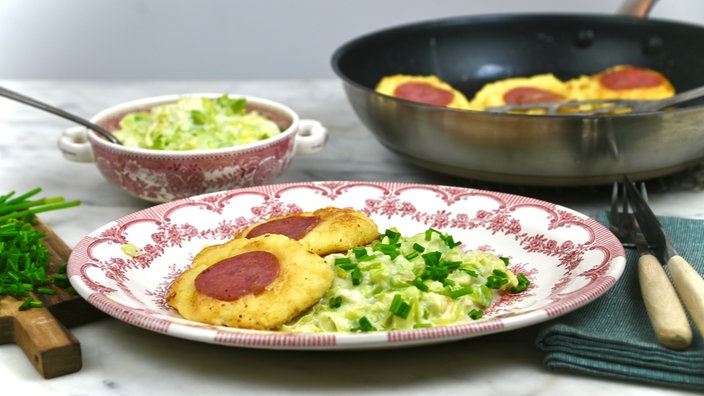 Ein angerichteter Teller mit Klitschkes, daneben Lauchgemüse in Sauce. Im Hintergrund sieht man eine Schüssel mit dem restlichen Lauchgemüse und die Pfanne mit den restlichen Klitschkes. 