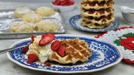 Eine Lütticher Waffel ist mit Sahne und Erdbeeren auf einem Teller angerichtet, im Hintergrund steht ein Teller mit weiteren Waffeln und ein Backblech mit Teig