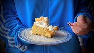 Ein Stück Rührkuchen mit Mandarinen ist auf einem Teller angerichtet, der von einer Frau mit blauer Jacke gehalten wird
