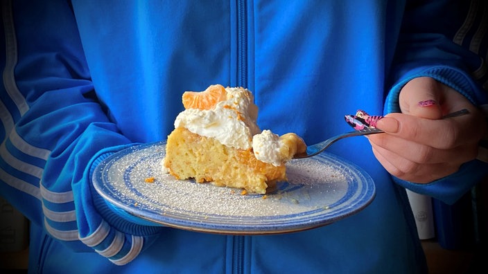 Ein Stück Rührkuchen mit Mandarinen ist auf einem Teller angerichtet, der von einer Frau mit blauer Jacke gehalten wird