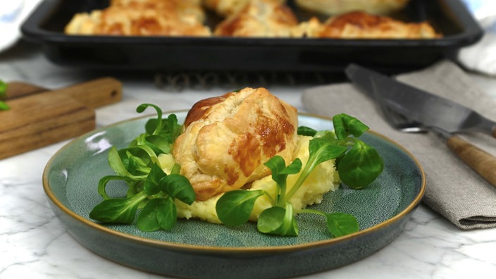Ein Sauerkrautpastetchen ist auf Kartoffelpüree und Feldsalat angerichtet. Um den Teller liegen eine Serviette mit Besteck, ein Holzbrettchen und im Hintergrund ein Blech mit weiteren Pasteten