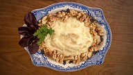 Von oben fotografiert: Eine große weiß-blau verzierte Schale mit Selleriepürree, garniert mit Walnüssen, auf einem braunen Holztisch. 