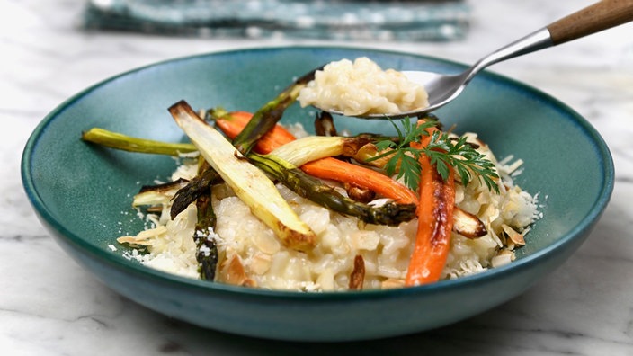 Das Risotto wird auf einem blauen Teller serviert. Darauf ist das geröstete Gemüse platziert. Mit einem Löffel wird ein wenig Risotto angehoben.
