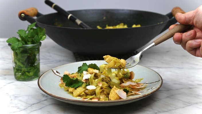 Spitzkohl und Hähnchen in Curry-Kokos-Sauce ist auf einem Teller angerichtet, im Hintergrund stehen ein Glas mit frischem Koriander und das restliche Curry in einer Wok-Pfanne